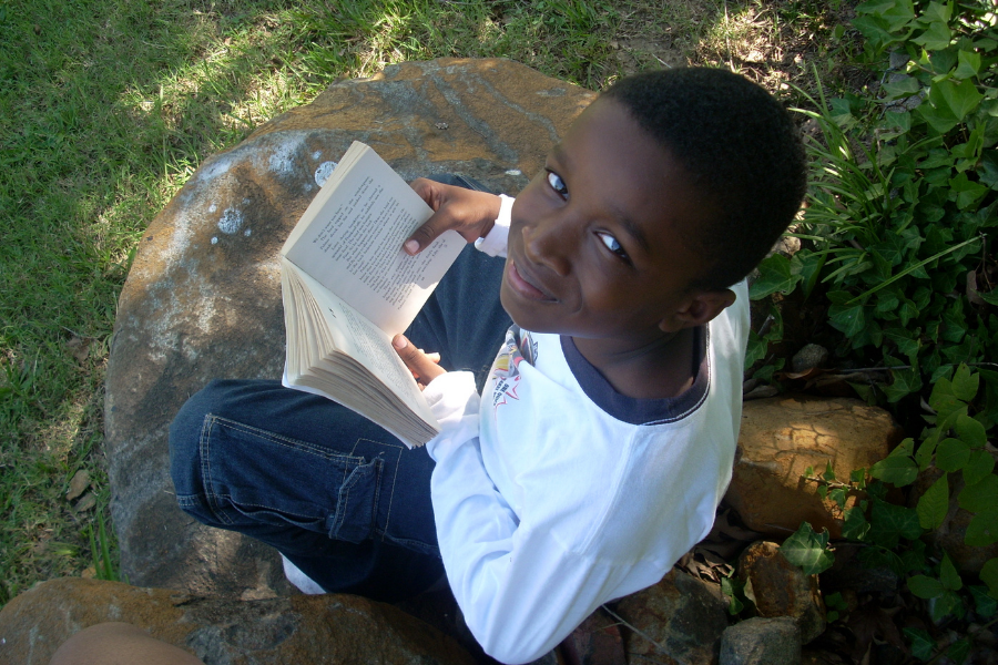A kid sits with a book and happily smiles at the camera, points to recommendations for kids page.