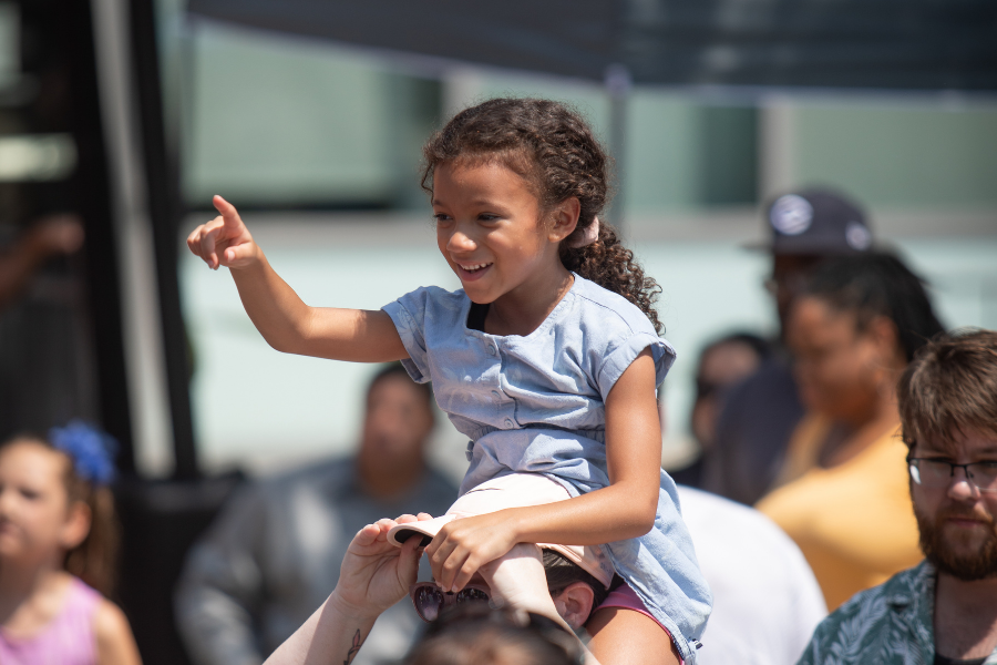 A young child sits on someone's shoulders and happily points.
